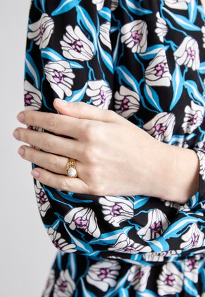 Schwarzer Stoff mit weißen Blumen und blauen Blättern; goldener Ring mit strukturiertem Mittelpunkt; Hand sanft auf einem gemusterten Ärmel ruhend.