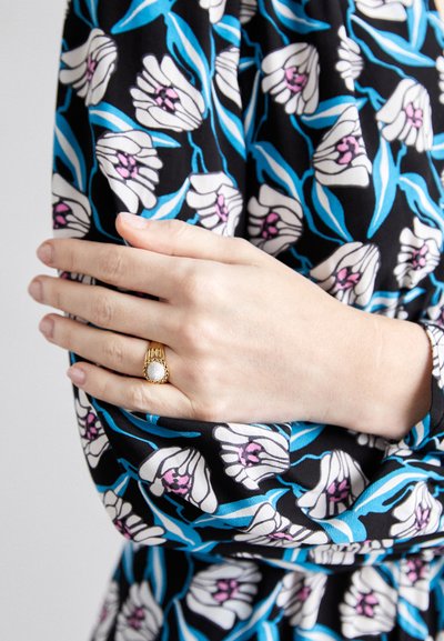 Schwarzer Stoff mit weißen Blumen und blauen Blättern; goldener Ring mit strukturiertem Mittelpunkt; Hand sanft auf einem gemusterten Ärmel ruhend.