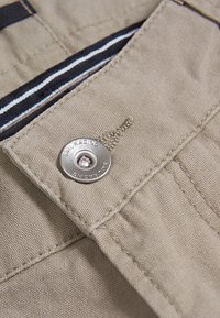 Tejido beige con un acabado texturizado presenta un botón de metal plateado embossado con "SAIL RACING". Incluye un forro interior a rayas.
