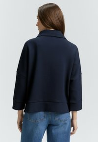 Mujer con cabello largo castaño, vistiendo una chaqueta suelta azul marino y jeans azules, vista desde atrás contra un fondo liso.