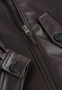 Mørkebrun læderjakke tæt på med metallynlås, knappet klaplomme og manchet med sorte knapper og detaljeret syning.