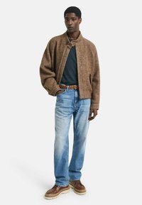 Veste en chevrons marron avec un col haut, associée à un t-shirt sombre, un jean bleu clair et des chaussures marron à semelle épaisse.