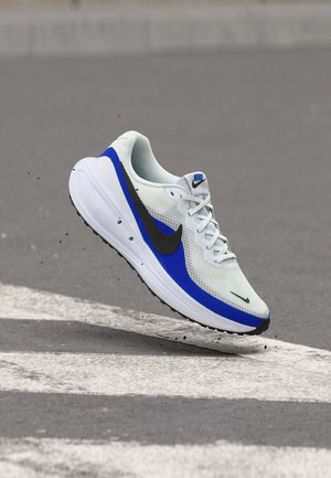 Zapatilla de running Nike blanca y azul con el logotipo negro que flota sobre una carretera asfaltada con líneas blancas de paso de peatones.