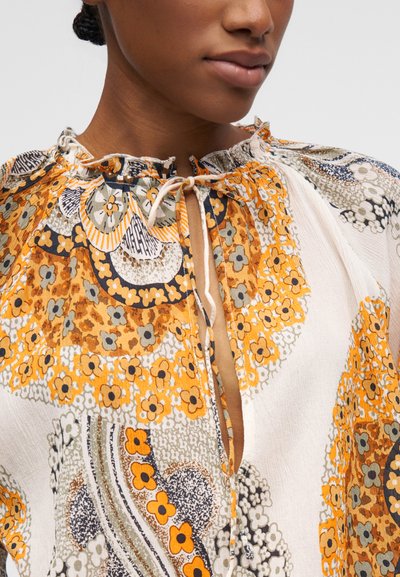 Femme portant un chemisier à motifs floraux jaune moutarde et blanc avec un col à volants noué à l'avant.
