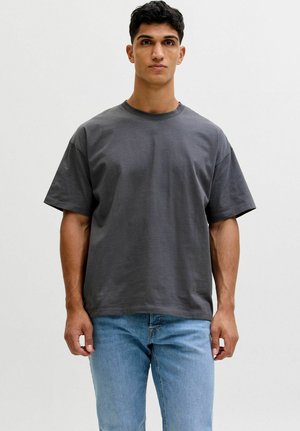 Giovane uomo in piedi, con indosso una maglietta oversize grigio scuro tinta unita e jeans azzurri chiari, su sfondo bianco.