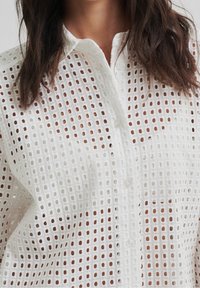 White, button-up shirt featuring a textured open weave pattern with small circular cutouts. Soft fabric with a collar and long sleeves.