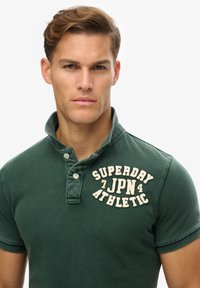 Groene polo met korte mouwen, gestructureerde stof, knoopkraag en een crèmekleurig geborduurd logo met de tekst "SUPERDRY JPN ATHLETIC."