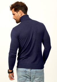 Marineblauer, schmal geschnittener Strickpullover mit hohem Kragen, gerippten Bündchen und Saum; getragen mit hellblauen Jeans, von hinten betrachtet.