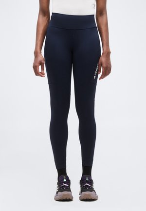 Kvinde iført sorte træningsleggings med sidelommer og sorte og lilla løbesko, stående mod en ensfarvet baggrund.