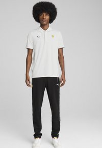 Hvid polo-shirt med Ferrari-logo, parret med sorte sweatpants med sideløsning. Afsluttet med hvide træningssko.