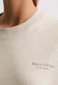 Zbliżenie na osobę w beżowej koszulce z okrągłym dekoltem, z napisem "Marc O'Polo EST. W STOCKHOLM" na piersi.