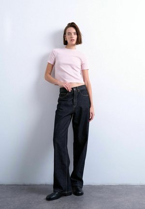 Woman wearing light pink short-sleeve cropped top, loose black jeans, and black shoes, standing against a plain white wall.