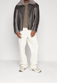 Mørkebrun læderjakke med grå shearling-detaljer, lynlås foran, kombineret med en tekstureret brun sweater og hvide bukser med vide ben.