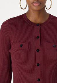 Burgundi bordás kardigán kerek nyakkivágással, fekete gombokkal és két elülső zsebbel. Simaszerű textúrája függőleges csíkokkal.