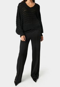 Schwarzer Strickpullover mit V-Ausschnitt, der horizontale Streifen und eine lockere Passform aufweist. Kombiniert mit hochgeschnittenen schwarzen Hosen und spitzen schwarzen Absätzen.