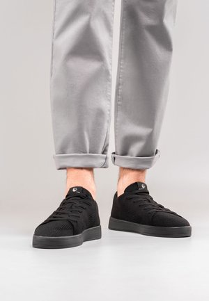 Black knit sneakers with lace-up design, featuring a textured upper and thick rubber sole. Paired with gray cuffed trousers.