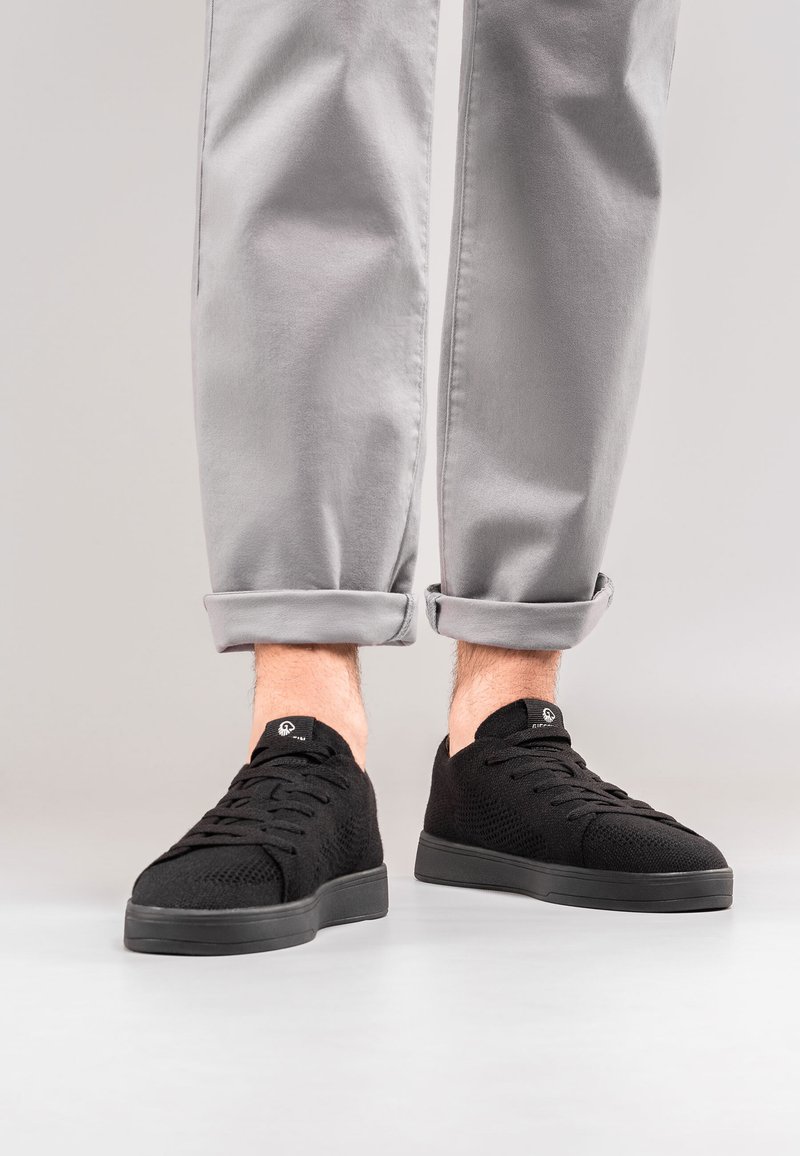 Czarne dziane sneakersy z sznurowaną konstrukcją, z teksturowaną cholewką i grubą gumową podeszwą. W zestawie z szarymi spodniami z mankietami.