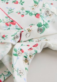 Tessuto bianco con motivo floreale rosso e verde che mostra bottoni a pressione metallici lungo un bordo curvo.