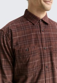 Chemise à carreaux brune en tissu texturé, dotée d'un col à boutons, de deux poches avant et de boutons bruns.