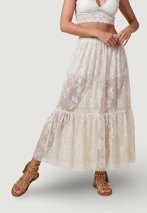 Donna che indossa un bralette in pizzo bianco e una lunga gonna di pizzo trasparente abbinati a sandali gladiatore marroni con borchie, in piedi con le gambe divaricate.