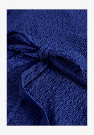 Tamsiai mėlynos spalvos tekstūruota medžiaga su surišta kaspino formos detalė tuo pačiu iš tos pačios medžiagos ir spalvos, išryškinanti medžiagos tekstūrą ir mazgo formą.