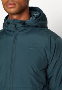 Homme portant une veste à capuche bleu sarcelle foncé avec fermeture éclair et logo Nike sur la poitrine, visible des épaules aux lèvres sur fond blanc.