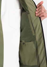 Vnitřek olivově zelené bundy s měkkou podšívkou, který obsahuje náprsní kapsu zabezpečenou patentním knoflíkem a zipové uzavření podél okraje.