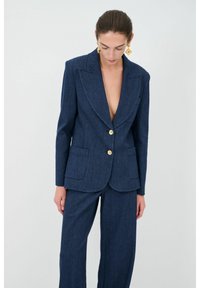 Blazer in denim di un blu intenso con revers a scollo a V, chiusura con un singolo bottone dorato e doppie tasche frontali. Vestibilità sartoriale con pantaloni coordinati.