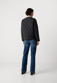 Donna con capelli corti e ricci, che indossa un giubbotto nero trapuntato, jeans blu e stivali con tacco, in piedi di spalle contro uno sfondo semplice.