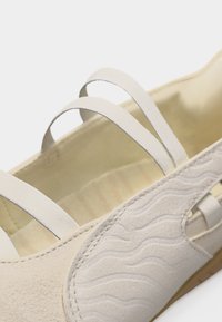 Chaussure beige à enfiler avec motif ondulé texturé sur le côté et trois bandes élastiques sur le dessus pour un ajustement sûr.