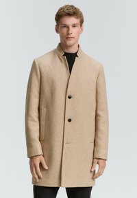 Beige wollen jas met een rechte snit, hoge kraag en twee voorzakken. Sluit met drie zwarte knopen. Gekocht over een zwarte trui.