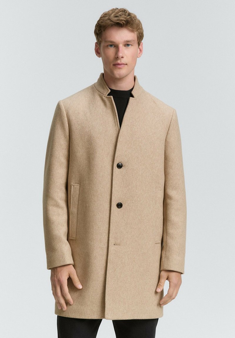 Beige wollen jas met een rechte snit, hoge kraag en twee voorzakken. Sluit met drie zwarte knopen. Gekocht over een zwarte trui.