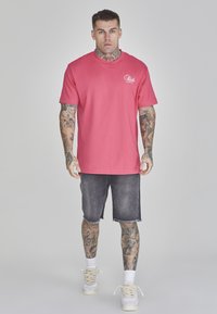 Camiseta de manga corta rosa con cuello redondo y logo blanco, combinada con pantalones cortos de mezclilla oscura y zapatillas blancas, con tatuajes detallados en los brazos.