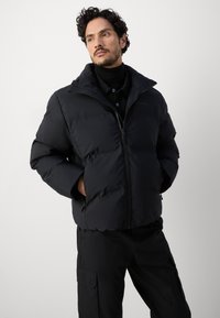 Veste rembourrée noire avec col montant et fermeture éclair à l'avant. Comprend des poches latérales et une finition lisse et mate. Portée par-dessus une chemise noire.