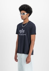 Sort bomulds t-shirt med et centralt gråt logo. Korte ærmer, parret med lyseblå bukser. Accessories inkluderer en kædehalskæde og solbriller.