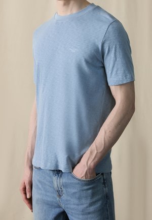 Homme portant un t-shirt bleu clair à manches courtes et un jean bleu, une main dans la poche, sur un fond neutre.