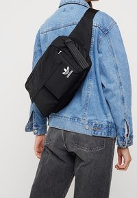Person som bär en svart Adidas crossbody-väska över en ljusblå denimjacka och mörkgrå jeans, vänd bort från kameran.