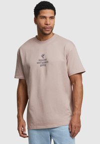 Camiseta de algodón de color rosa claro con mangas cortas, cuello redondo y un gráfico de texto impreso en gris oscuro, que representa un pájaro y la frase "inocente hasta que se demuestre lo contrario."