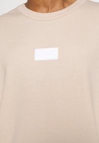 Light beige sweatshirt made from soft cotton, featuring a white embossed Adidas logo patch on the chest and a round neckline.