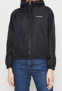 Chaqueta cortavientos negra con cremallera frontal, capucha, puños elásticos y dobladillo. Cuenta con un pequeño parche de logo en el pecho y dos bolsillos frontales.