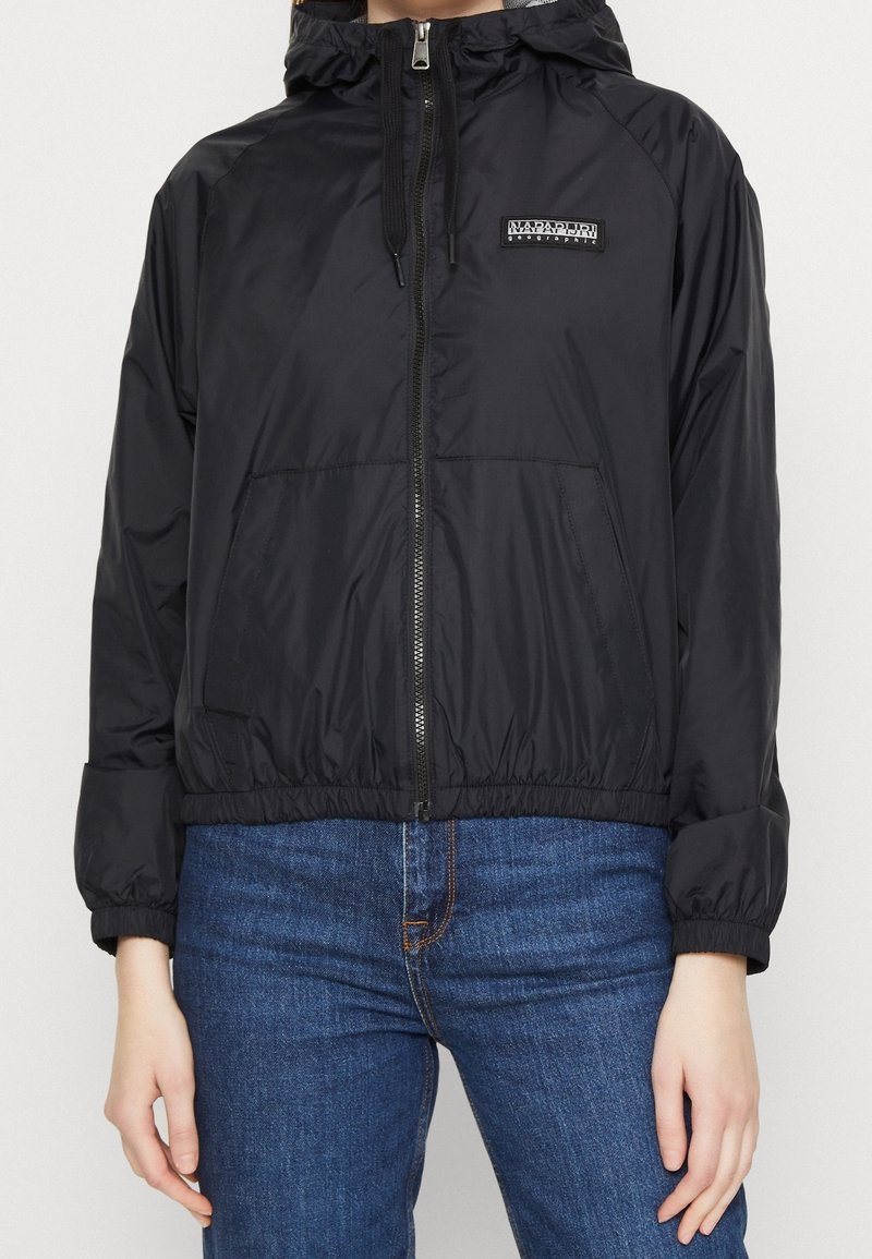 Chaqueta cortavientos negra con cremallera frontal, capucha, puños elásticos y dobladillo. Cuenta con un pequeño parche de logo en el pecho y dos bolsillos frontales.