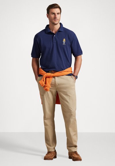 Jūras spēku polo krekls ar logo, savienots ar khaki biksēm. Oranžs džemperis sasietas ap vidukli un brūni apavi noslēdz apģērbu.