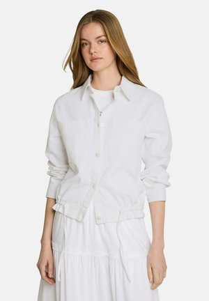 Jeune femme aux longs cheveux blonds portant une veste blanche à boutons avec cordons et une robe blanche sur un fond uni.