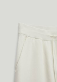 Section de pantalons de survêtement écru montrant une taille élastique avec cordon de serrage et une poche latérale sur un fond clair uni.