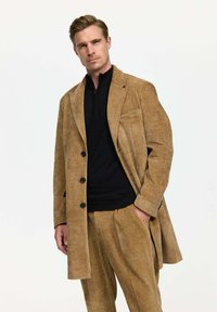 Blazer de pana color marrón con solapa de muesca, dos bolsillos frontales y botones negros, combinado con un suéter negro con cremallera y pantalones a juego.