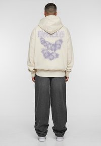 Beige hættetrøje med et stort lilla sommerfuglegrafik på ryggen, løs pasform, ribbede manchetter, og båret med grå vide jeans og hvide sneakers.