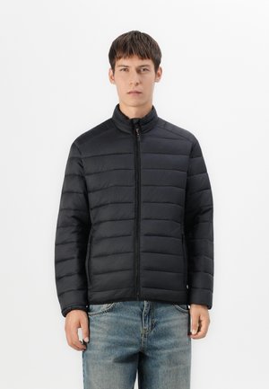 Jeune homme portant une veste noire matelassée zippée et un jean bleu, debout devant un fond blanc uni.