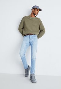 Sweter z dzianiny w oliwkowym kolorze o teksturowej fakturze, jasnoniebieskie dżinsy, szare buty sportowe i szara czapka. Prosty wygląd z luźnym krojem.