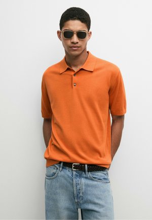Mann mit dunkler Sonnenbrille, orangefarbenem Kurzarm-Poloshirt, hellblauen Jeans und schwarzem geflochtenem Gürtel steht vor einem schlichten Hintergrund.