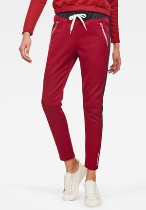 Persona con pantalones deportivos rojos con rayas negras laterales, cordón blanco, bolsillos con cremallera y zapatillas casuales blancas sin cordones.